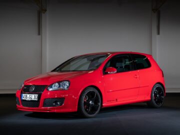 A red two-door hatchback Volkswagen Golf GTI with black alloy wheels, parked in an indoor area. The license plate reads 