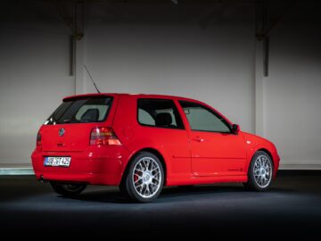 A red two-door compact Volkswagen Golf GTI is parked inside under bright lights. The vehicle has a sporty design and alloy wheels. The license plate reads 