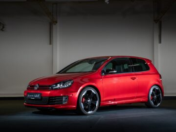 Ein roter Volkswagen Golf GTI steht im Innenraum auf einem dunklen Boden, wobei die Seite und die Vorderseite des Autos beleuchtet sind.