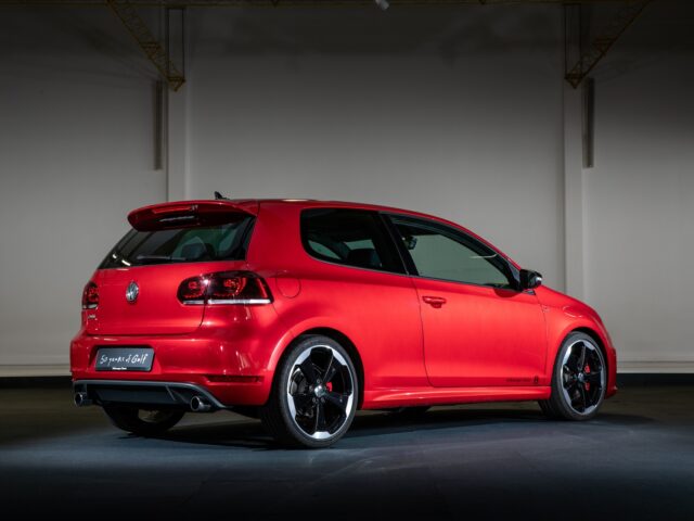 A red Volkswagen Golf GTI hatchback with a sporty design is parked inside, seen from the rear. The vehicle is equipped with black alloy wheels and has visible taillights and dual exhaust pipes.