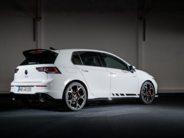 A white Volkswagen Golf GTI hatchback with black details is parked inside, seen from the left rear. The car features sporty red brake calipers and dual exhaust pipes.