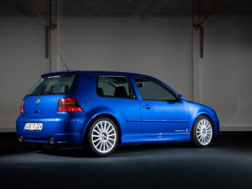 A blue two-door Volkswagen Golf GTI R32 is parked in an interior space with minimal lighting. The car, seen from the rear, features sporty design elements and alloy wheels.