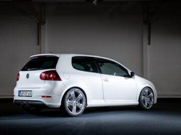 Inside, on a dark background is a white compact Volkswagen Golf GTI with a sleek design. The car has a glossy exterior and silver alloy wheels. The rear of the car, including the rear license plate, is visible.