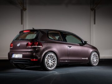Inside, a dark brown Volkswagen Golf GTI with European license plates is parked. The car has two doors and is illuminated by studio lights, highlighting its sleek design and alloy wheels.