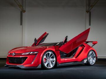 A sleek red sports car with open butterfly doors, with a futuristic design reminiscent of a Volkswagen Golf. The car has a low profile, angular lines and a prominent grille with a race-inspired emblem.