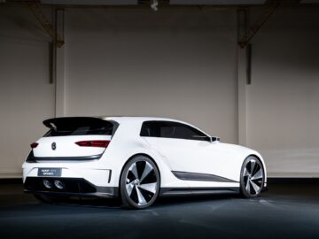 A white, sleek, futuristic concept car reminiscent of a Volkswagen Golf is displayed in a dimly lit showroom against a plain background.