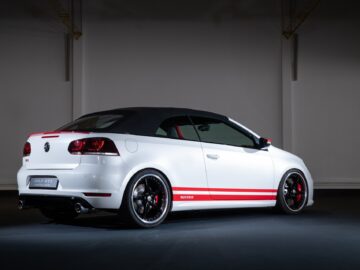 A white Volkswagen Golf GTI convertible with black soft top, red striping and custom rims is parked in a well-lit interior space.