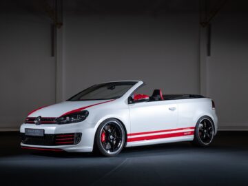 A white Volkswagen Golf GTI Cabriolet with red accents is displayed in a dark room. The car's roof is open, revealing a striking black and red interior.