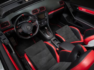 Interior of a modern black and red Volkswagen Golf GTI sports car with a leather steering wheel, touchscreen, manual transmission and sporty bucket seats with red accents.