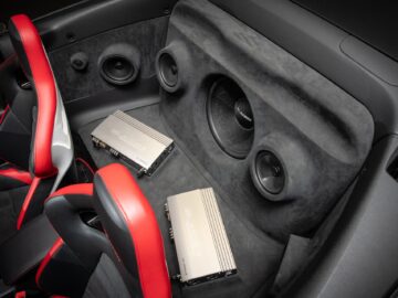 Het interieur van een Volkswagen Golf met rood en zwart lederen stoelen, met een geluidssysteem inclusief een subwoofer en twee autoradioversterkers die achter de stoelen zijn gemonteerd.