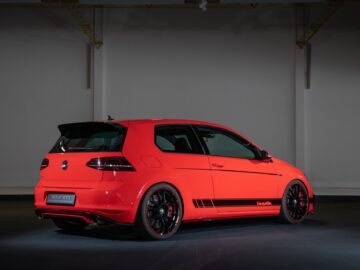 Ein roter Volkswagen Golf GTI mit schwarzen Felgen und einem Spoiler ist im Innenraum geparkt, von hinten links gesehen. Der Volkswagen Golf zeigt sportliche Designelemente vor einem dunklen, schattigen Hintergrund.