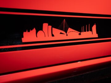 Rotes und schwarzes Bild, das die Silhouette einer Stadtsilhouette mit bemerkenswerten Gebäuden und Strukturen zeigt, während ein schnittiger Volkswagen Golf durch die Straßen fährt.