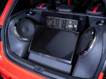 Das Bild zeigt den offenen Kofferraum eines roten Volkswagen Golf, der mit einem speziell angefertigten Soundsystem mit großen Lautsprechern, einem Verstärker und einem LCD-Bildschirm ausgestattet ist, die alle sauber installiert sind.