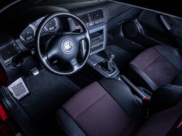 Interieur van een Volkswagen Golf-auto met een zwart stuur, dashboard met meters, handmatige versnellingspook en twee voorstoelen met een rood en zwart kleurenschema.