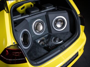 A Volkswagen Golf trunk with custom-installed audio equipment, with three large speakers integrated into a custom panel and a glass-covered compartment underneath containing electronic components.