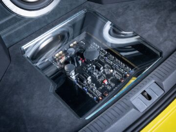 Close-up of an audio amplifier mounted under a glass cover in the trunk of a Volkswagen Golf, with visible electronic components and wiring.