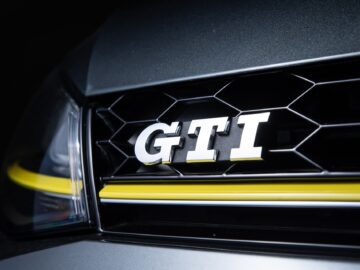 Close-up van een Volkswagen Golf-grille met het GTI-embleem, met een honingraatpatroon en gele accentstreep, een deel van de voorkant van het voertuig.