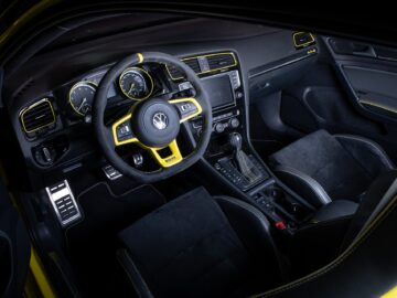 Interieur van een Volkswagen Golf gericht op de bestuurdersstoel, met een zwart en geel stuur, dashboard en diverse bedieningselementen. De bekleding is zwart met gele accenten.