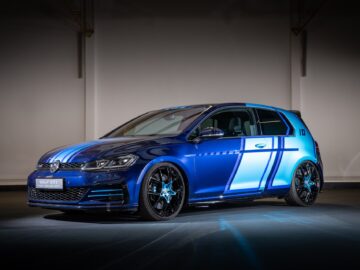 A blue Volkswagen Golf GTI with a sporty livery and upgraded wheels is parked inside against a plainly lit backdrop, showing off the iconic Volkswagen design.