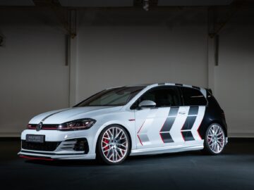 A white Volkswagen Golf GTI with custom black and gray emblems on the side, red accents and large alloy wheels is parked inside under artificial lights.