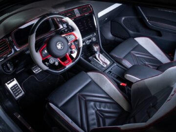 Driver's seat view of a Volkswagen Golf interior with black and red leather seats, a touchscreen dashboard, a steering wheel with the Volkswagen logo and a center console with various controls.