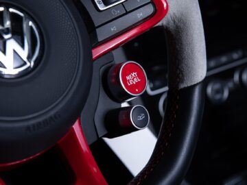 Close-up of a Volkswagen Golf steering wheel with the VW logo. A red button with the inscription 