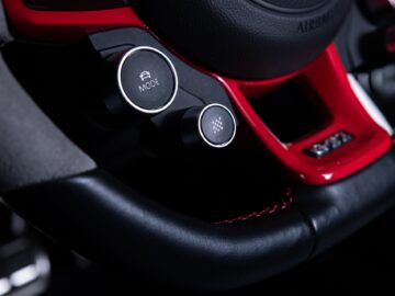 Close-up image of a Volkswagen Golf steering wheel with mode and control buttons, black leather with red stitching and part of the sleek dashboard design.