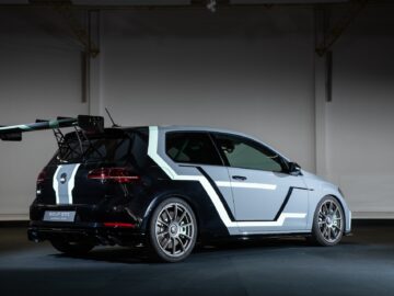 A customized Volkswagen Golf with geometric white and light blue designs on a gray body, fitted with a large rear spoiler and sporty alloy wheels, parked in a garage with subdued lighting.