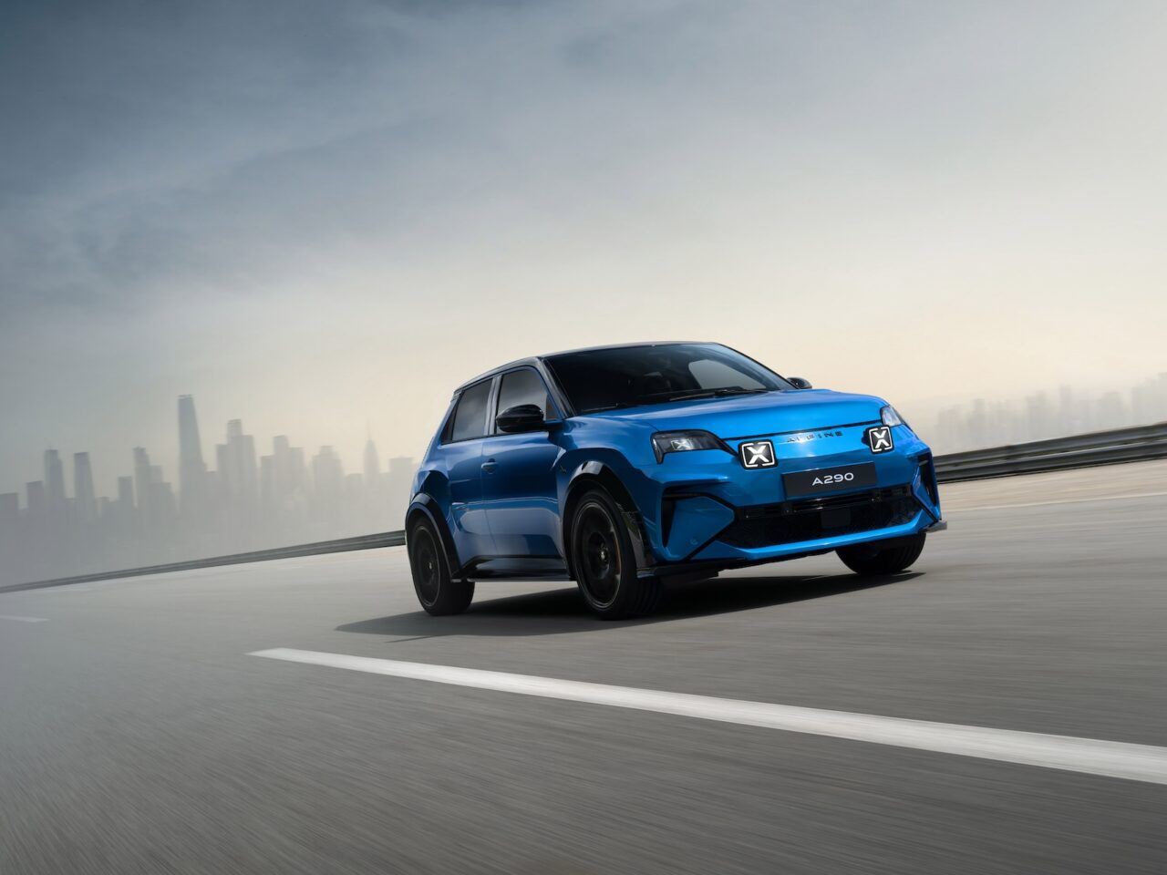 Een Alpine A290 met het opschrift "A290" rijdt over een brede weg met de skyline van een stad op de achtergrond onder een heldere hemel en laat zijn strakke ontwerp zien als een elektrische hot hatchback.
