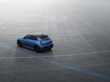 Een blauwe compacte SUV met een zwart dak, dat doet denken aan de stijlvolle Renault 5, staat geparkeerd op een groot, leeg asfaltoppervlak gemarkeerd met concentrische cirkellijnen.