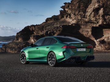 Un sedán de lujo verde está aparcado en un paisaje costero rocoso. El coche tiene un diseño elegante con cristales tintados. El océano y los acantilados se ven al fondo bajo un cielo despejado.