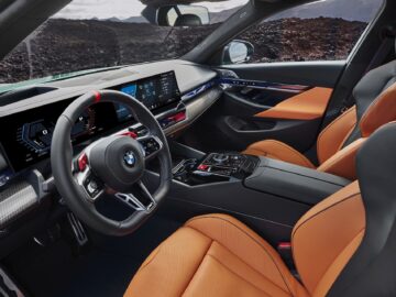 Interior view of a car with a sleek cockpit design, featuring a steering wheel with BMW logo, two digital displays and orange leather seats with black accents. The car stands against a rugged exterior background.