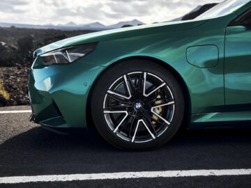 Close-up of the left front of a green BMW car, focusing on the steering wheel and part of the headlight, parked on an asphalt road with a rocky landscape in the background.