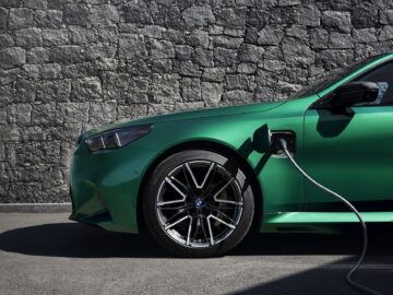 In profile, a green car is shown connected to an electric charging station, parked in front of a textured stone wall.