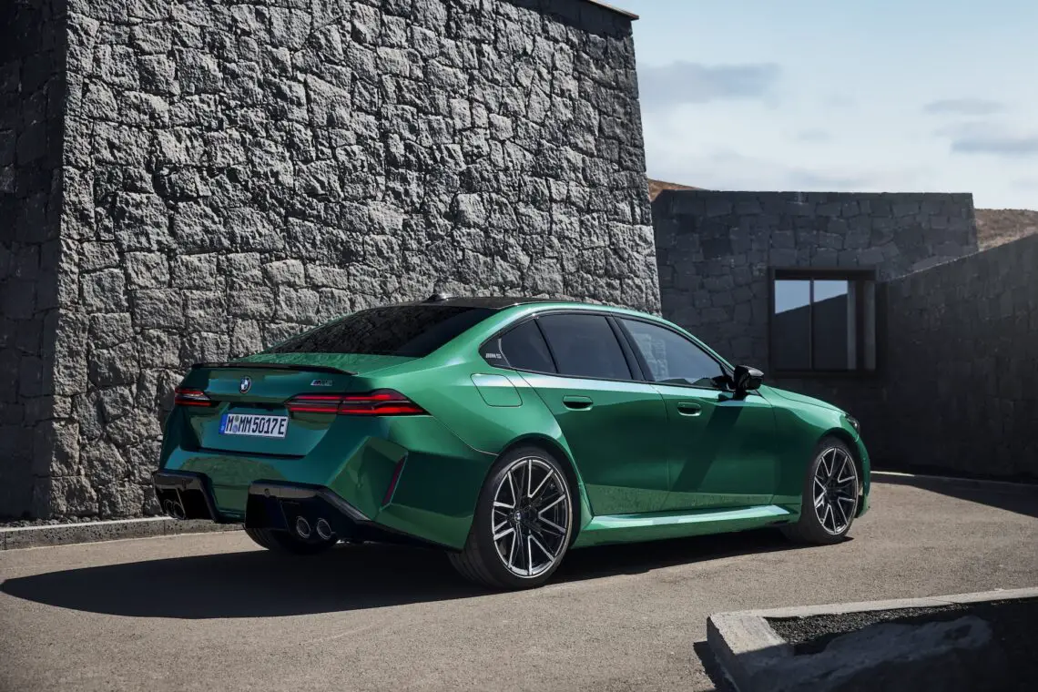 Een groene BMW M5 sedan staat geparkeerd op een verhard terrein voor stenen constructies, met een heldere hemel op de achtergrond.