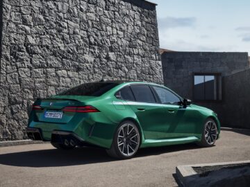 Een groene BMW M5 sedan staat geparkeerd op een verhard terrein voor stenen constructies, met een heldere hemel op de achtergrond.