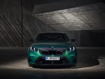 A green BMW car is parked in a dimly lit, angular concrete space, with its headlights on. The front view of the car shows a modern, sleek design.