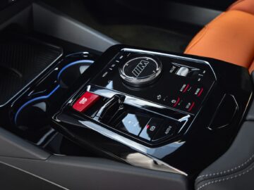 Close-up of the center console of a car with several control buttons, a large dial, cup holders and an orange seat visible in the background.