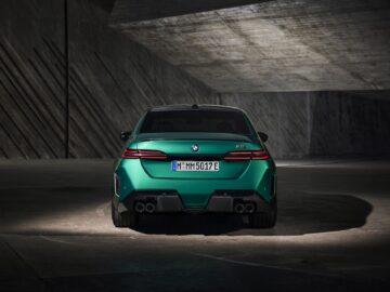 Achteraanzicht van een groene BMW M3 sedan in een slecht verlichte, industriële omgeving met betonnen muren en vloeren. De achterlichten en het kenteken van de auto zijn zichtbaar.