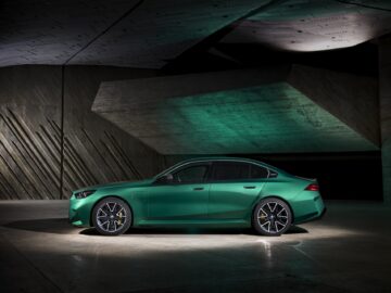 A green sedan with a sleek design and alloy rims is parked inside against an angular concrete wall, illuminated by overhead lights.