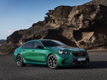 Un sedán verde está aparcado en un terreno rocoso bajo un cielo despejado. El coche tiene líneas limpias y rasgos de diseño moderno, con un fondo montañoso que proporciona contraste.