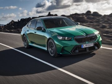 A green BMW sedan drives on a road with a rocky landscape and cloudy sky in the background.