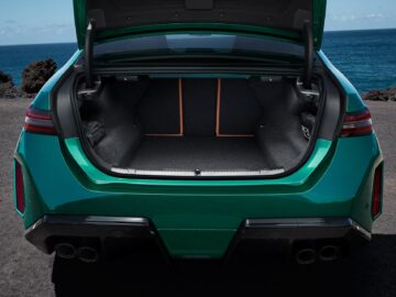 Green car with open trunk, revealing a spacious cargo area. The car is parked near the ocean on a clear day, with visible black rocky terrain and a blue sky in the background.