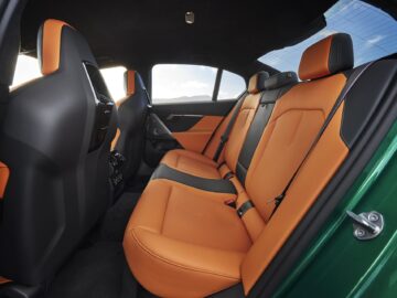 De afbeelding toont het achterste interieur van een auto met zwart-oranje lederen stoelen en zwarte bekleding, gezien door de open zijdeur tegen een buitenachtergrond.