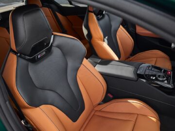 Interieur van een auto met voorstoelen bekleed met oranje en zwart leer met een sportief design en een 'M'-logo op de hoofdsteunen. De middenconsole bevat moderne bedieningselementen en een versnellingspook.