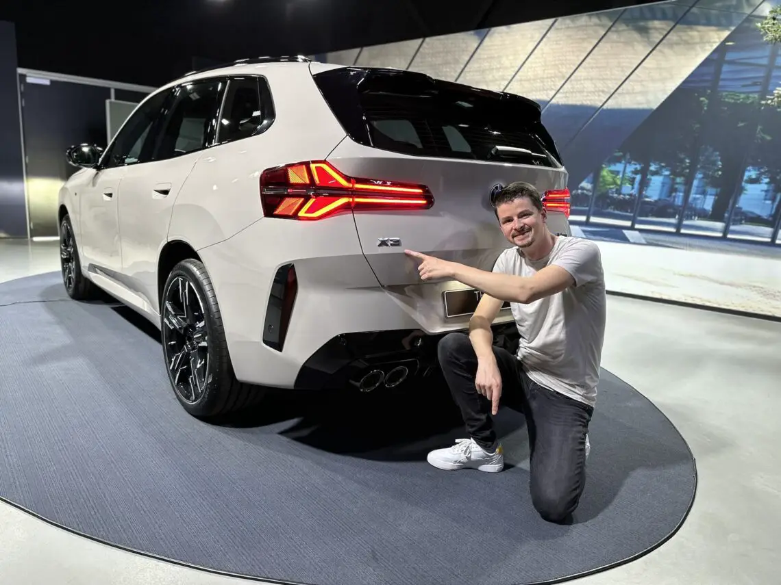Ein Mann kniet neben einem weißen BMW X3 2024 in einem Ausstellungsraum und deutet auf die Plakette auf der Rückseite des Fahrzeugs.