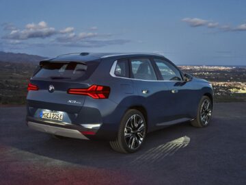 Une BMW X3 2024 bleue garée sur une colline surplombant une ville au crépuscule, avec les feux arrière allumés et la plaque d'immatriculation visible, offre une expérience de promenade étonnante.