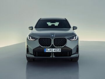 Frontansicht eines grauen BMW X3 (2024) SUV mit markantem Nierengrill, eingeschalteten Scheinwerfern und deutscher Plakette mit der Aufschrift 
