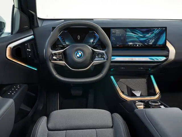 Innenansicht des neuen BMW X3 von 2024 mit futuristischem Lenkrad, digitaler Instrumententafel, großem Touchscreen-Display und blau beleuchteten Akzenten auf dem Armaturenbrett und der Konsole.