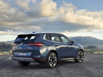 Une nouvelle BMW X3 SUV bleue est garée sur une plaine ouverte avec un paysage montagneux en arrière-plan. Le véhicule de 2024 porte un badge 